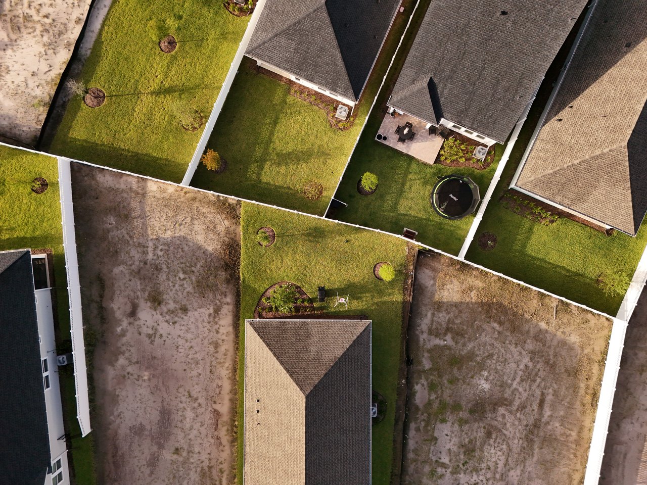 Aerial view of fenced residential yards, neighboring homes, vacant lots, and a backyard trampoline.