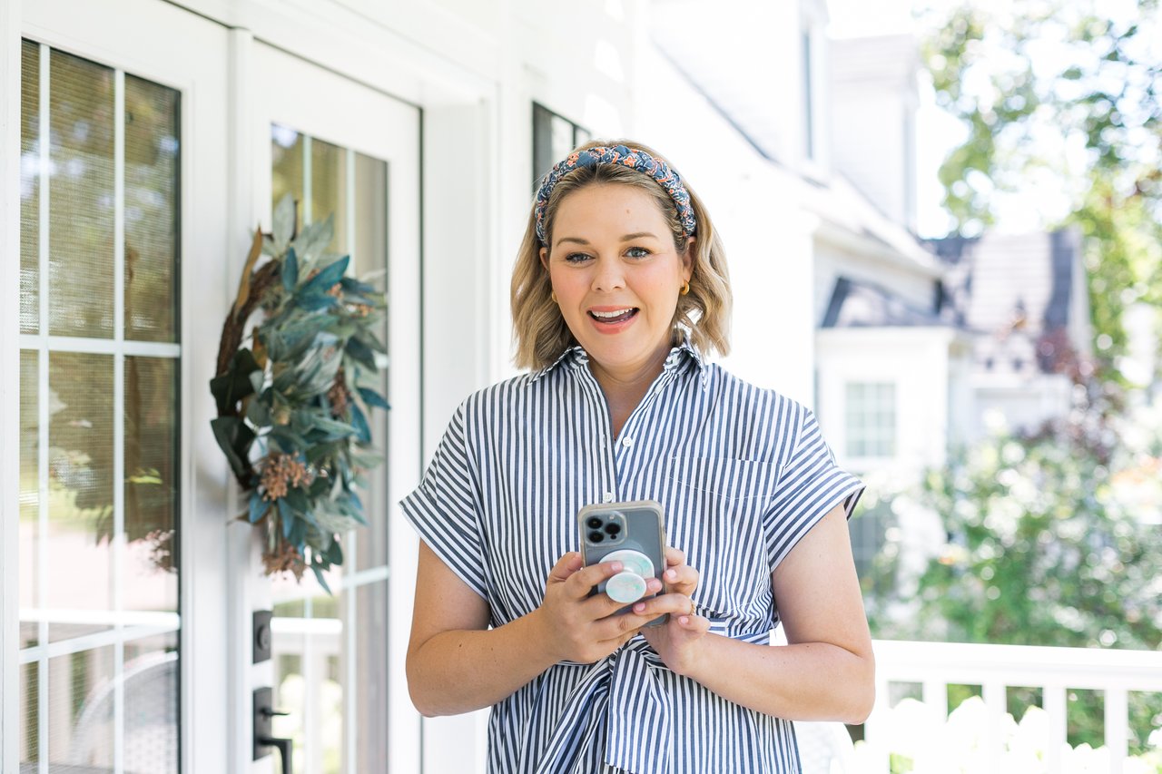 Madison WI real estate advisor Cait Berry standing on a welcoming front porch with her phone, sharing realistic tips on how to prepare your house for buyer showings.