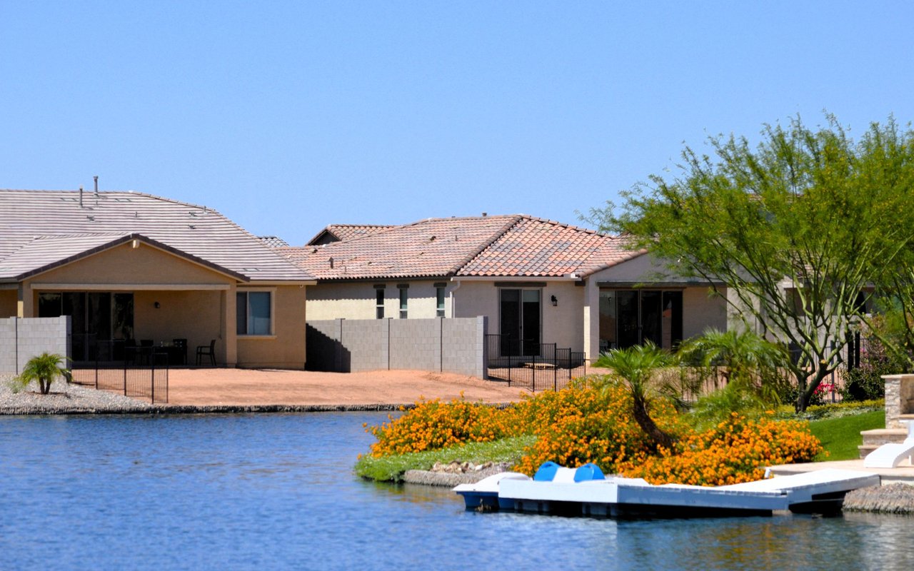 Stucco homes along a lake, with private docks and orange flowering shrubs on the shoreline.