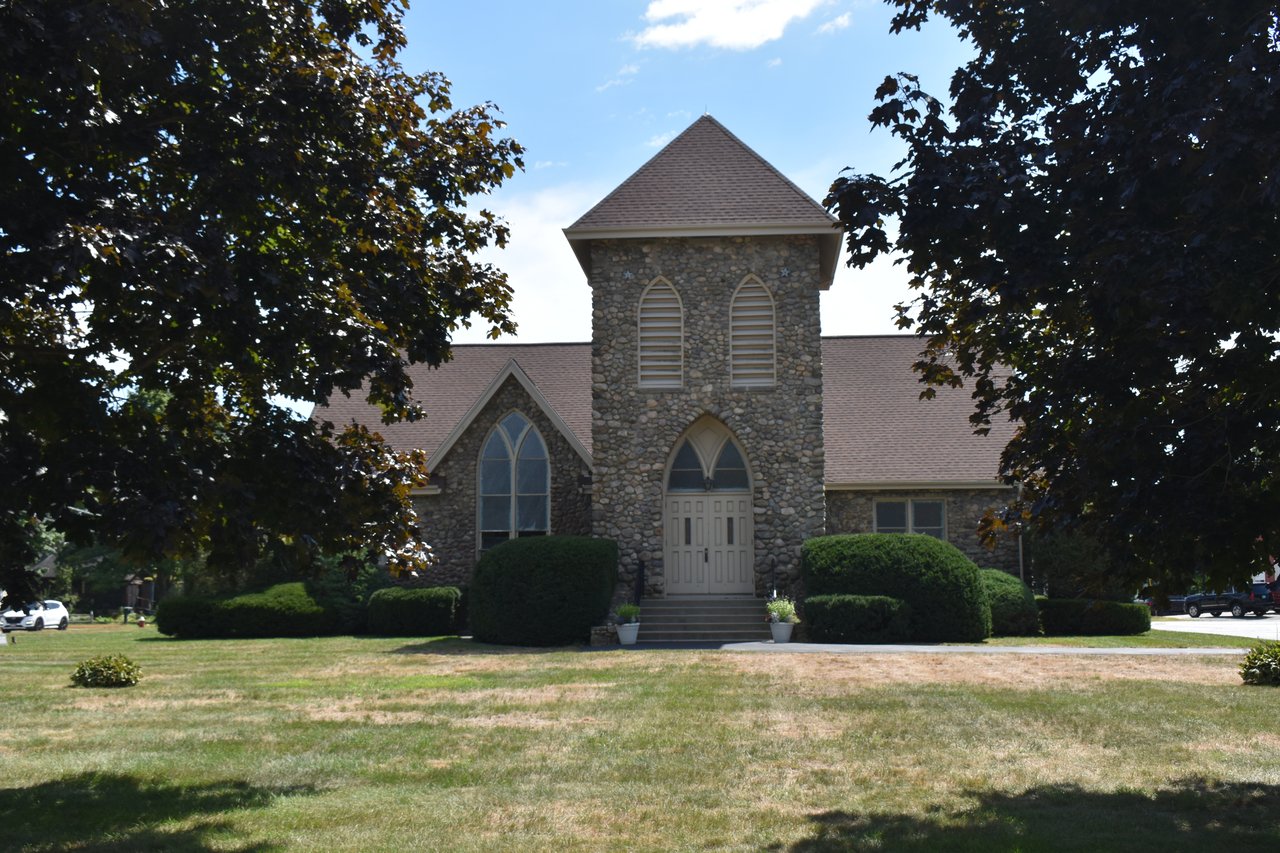 Stone church-like building with a central tower and pointed-arch windows, framed by trees across a lawn.