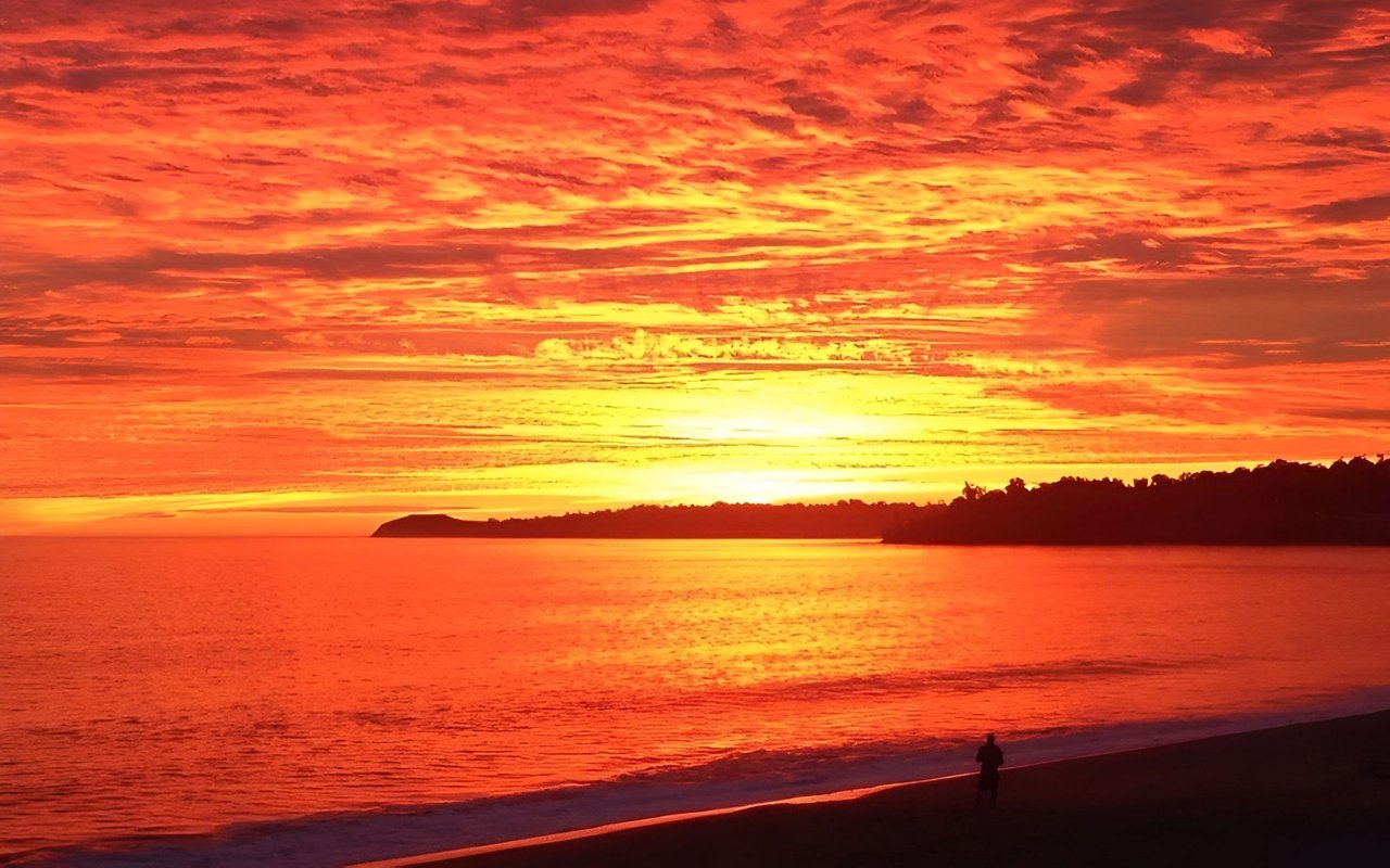 Orange-red sunset over the ocean, with a dark headland and a lone figure on the beach.