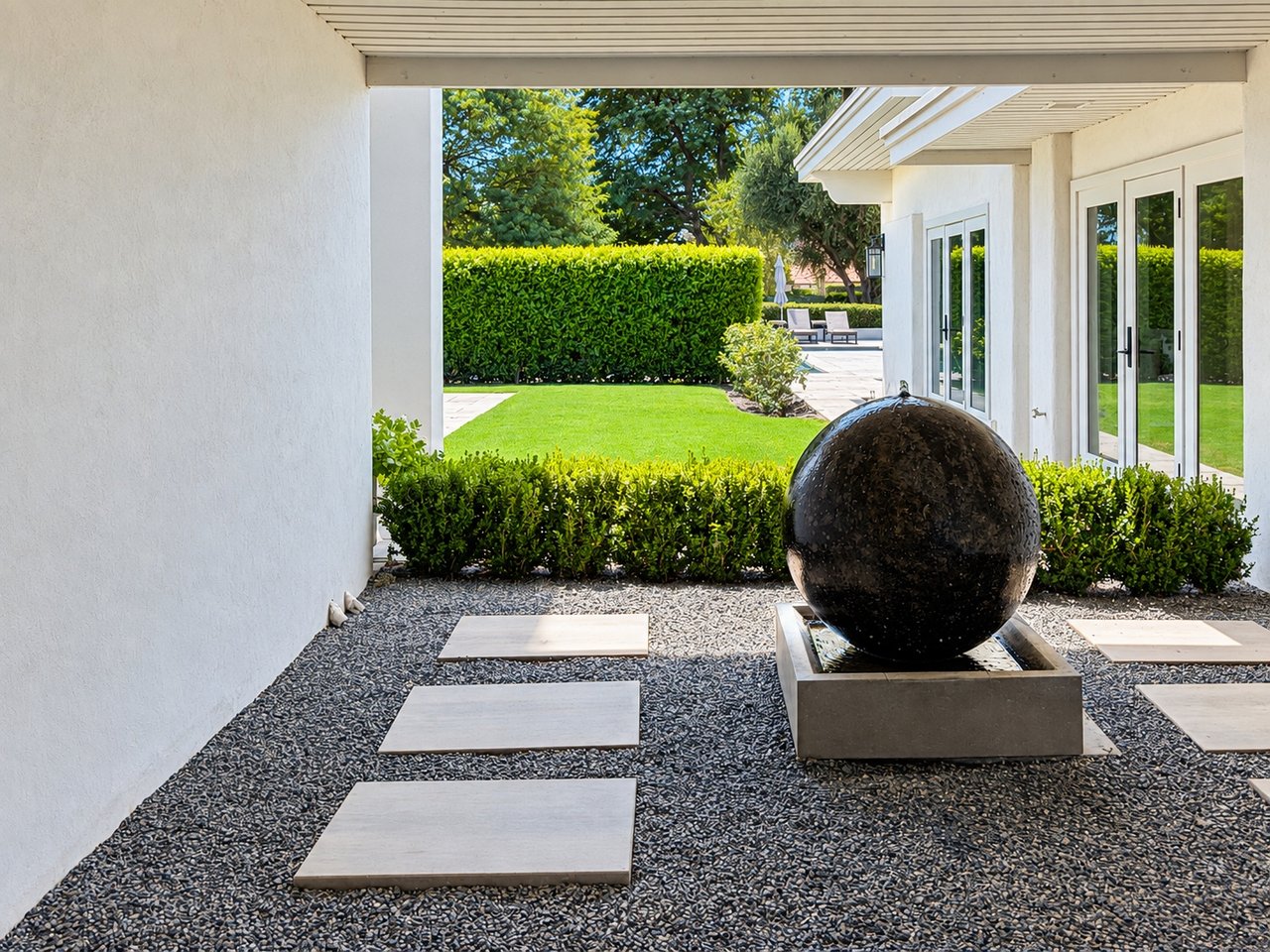 Modern home courtyard with a dark spherical fountain, gravel paths, clipped hedges, and a green lawn.