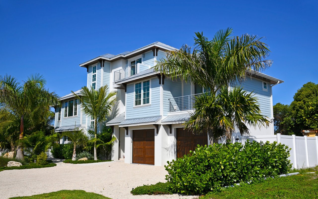Pale blue two-story house with metal roofs, palm trees, and a gravel driveway.