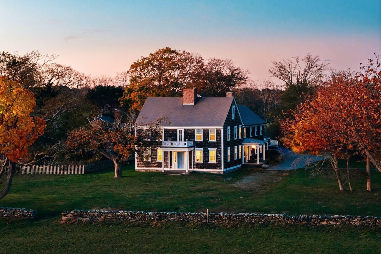 Dark-shingled two-story house with white trim among autumn trees at sunset.
