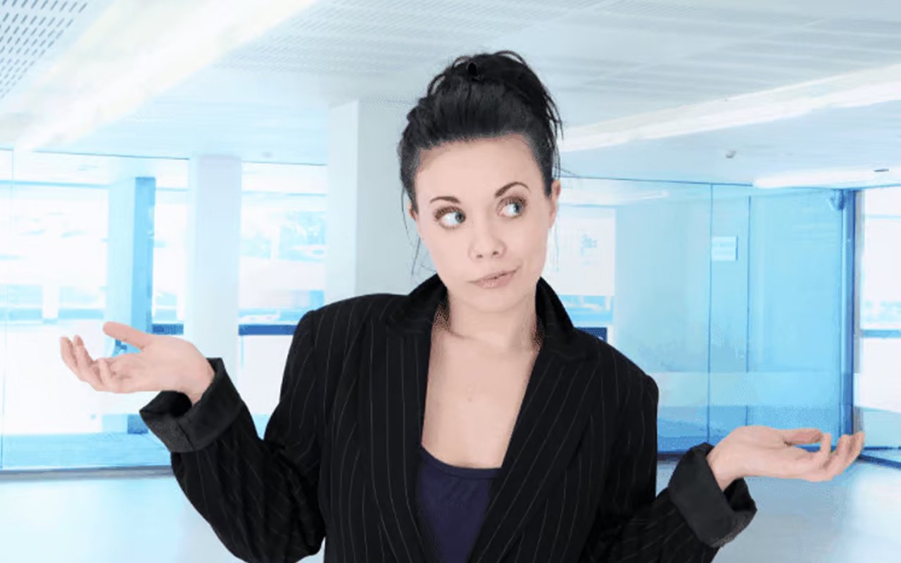 Woman in a pinstripe blazer shrugging with both hands raised in an office.