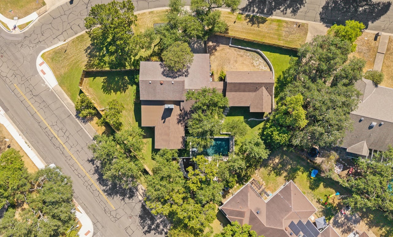 Aerial view of a single-story home with mature trees, a green lawn, and a backyard pool.