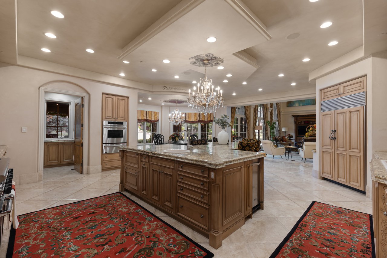 Luxury kitchen with a large wood island, granite counters, and ornate chandeliers opening to a living room.