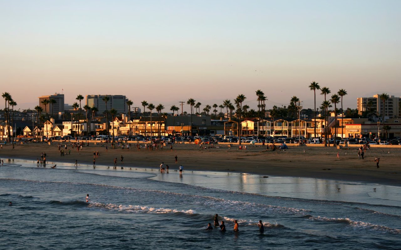 A view of a sunset at Newport Beach with people playing in the water, buildings and palm trees lining the shore.