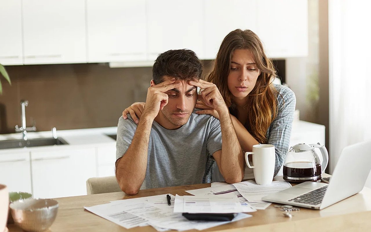 A stressed couple reviews paperwork at a kitchen table, with a laptop and coffee nearby.