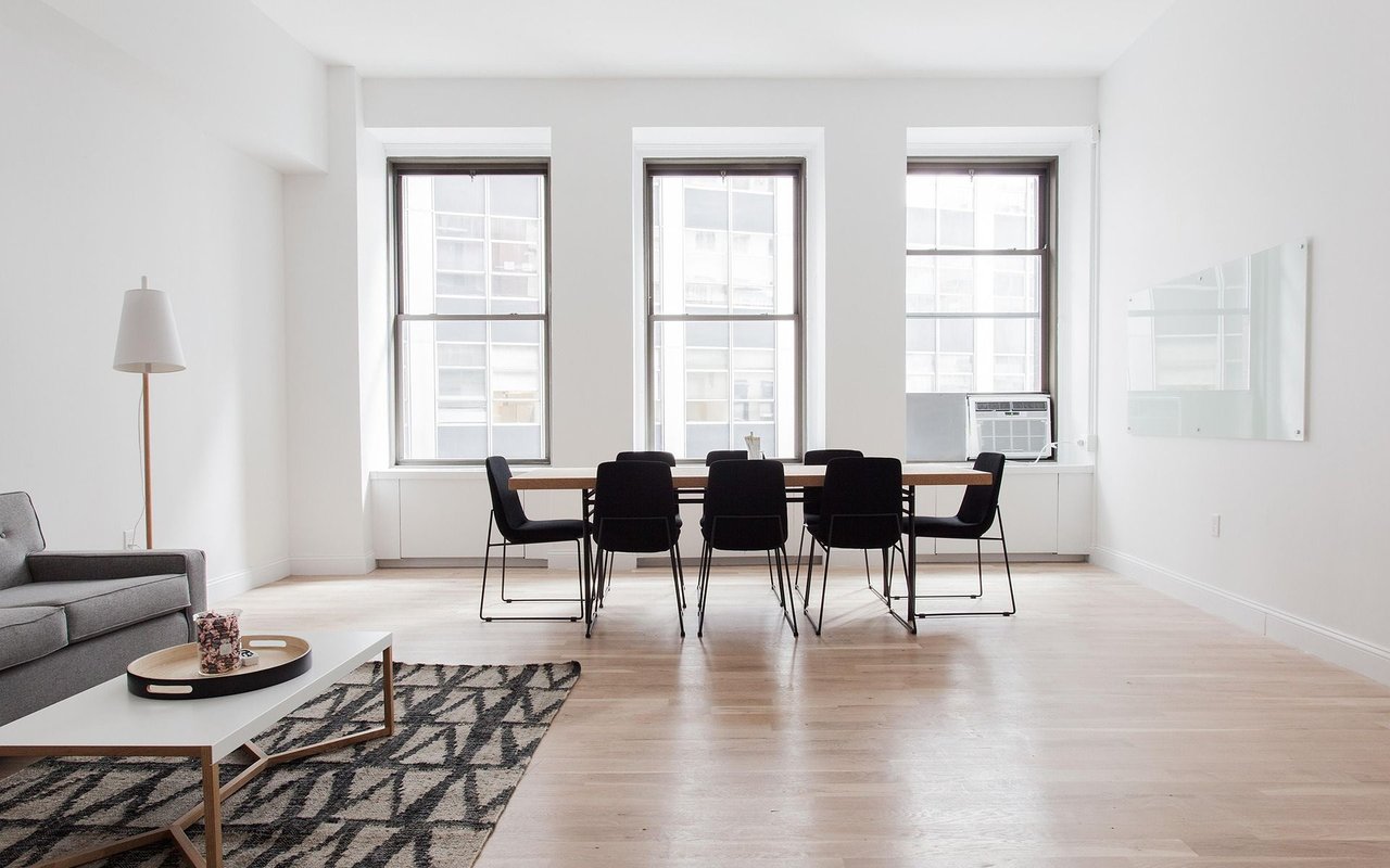 Bright open-plan living and dining room with three tall windows, pale wood floors, and black dining chairs.