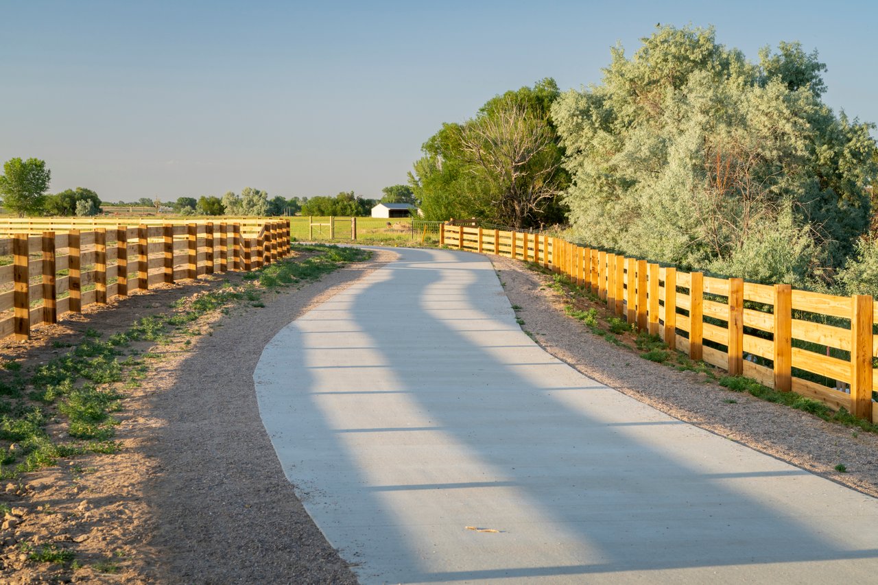 Paved walking path between wooden fences, with open fields and leafy trees.