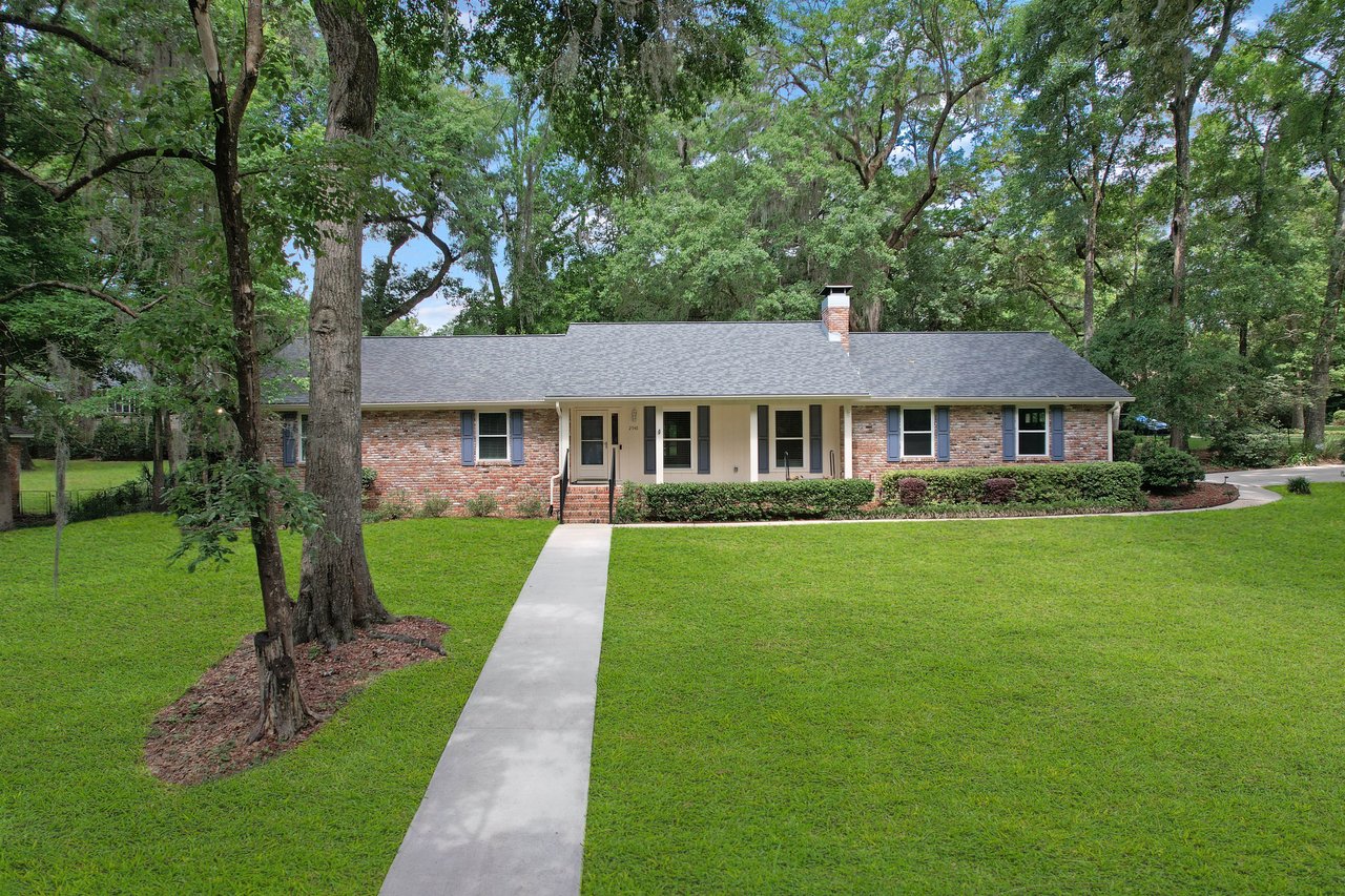 A charming brick ranch-style house with a gray roof is set against a lush backdrop of tall trees. A walkway leads to the porch, surrounded by a manicured lawn.