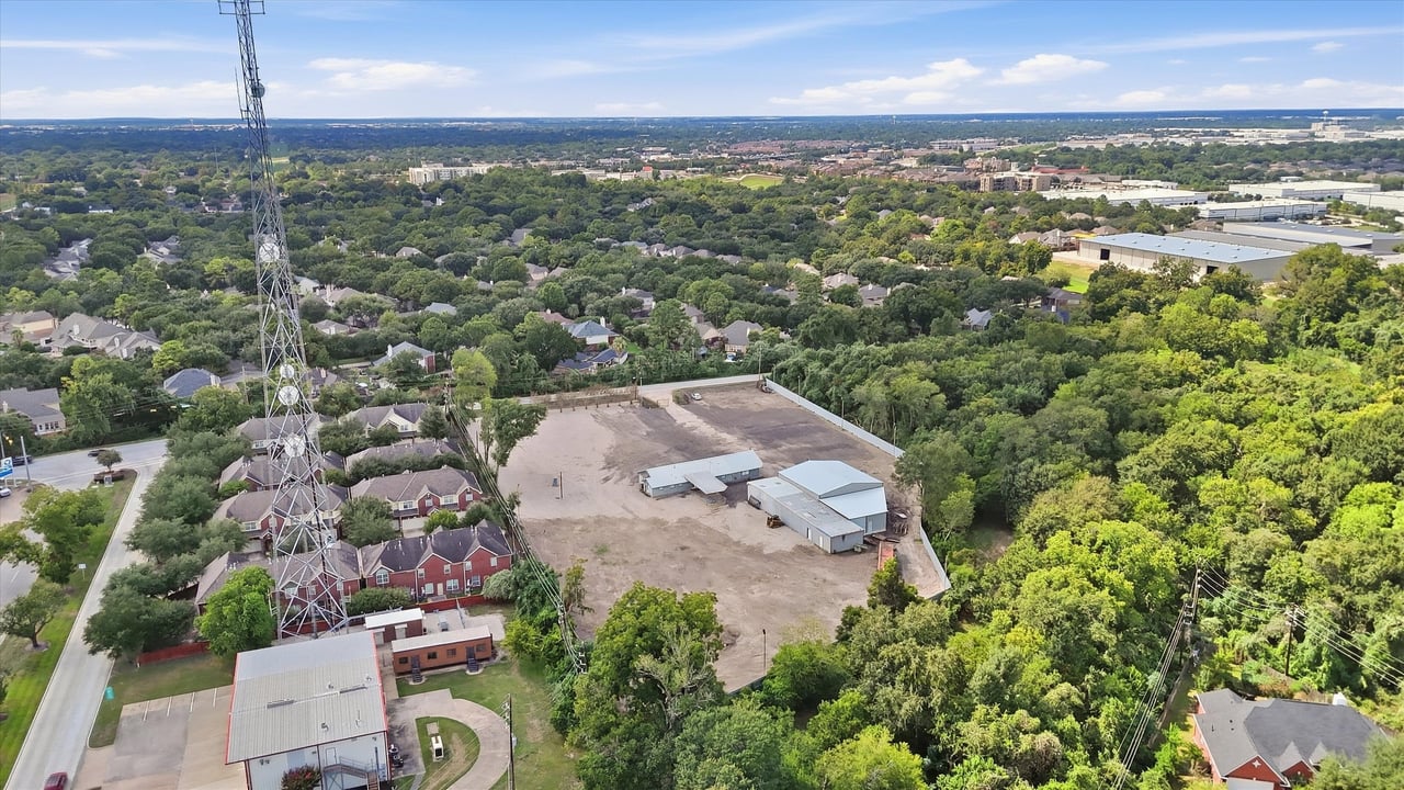 Aerial view of a cleared lot with metal buildings, surrounded by trees and homes.