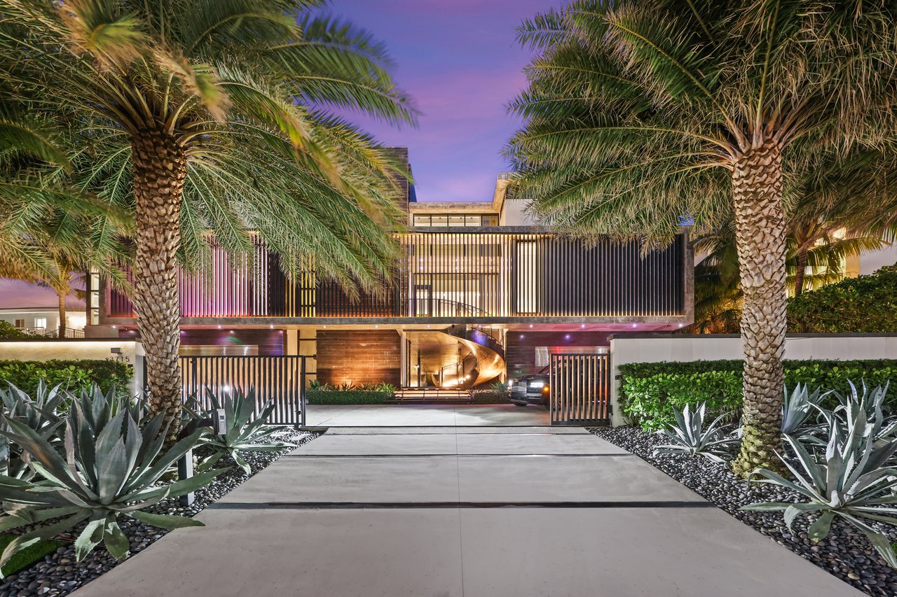 Modern luxury home with an illuminated facade, gated driveway, and palm trees at dusk.