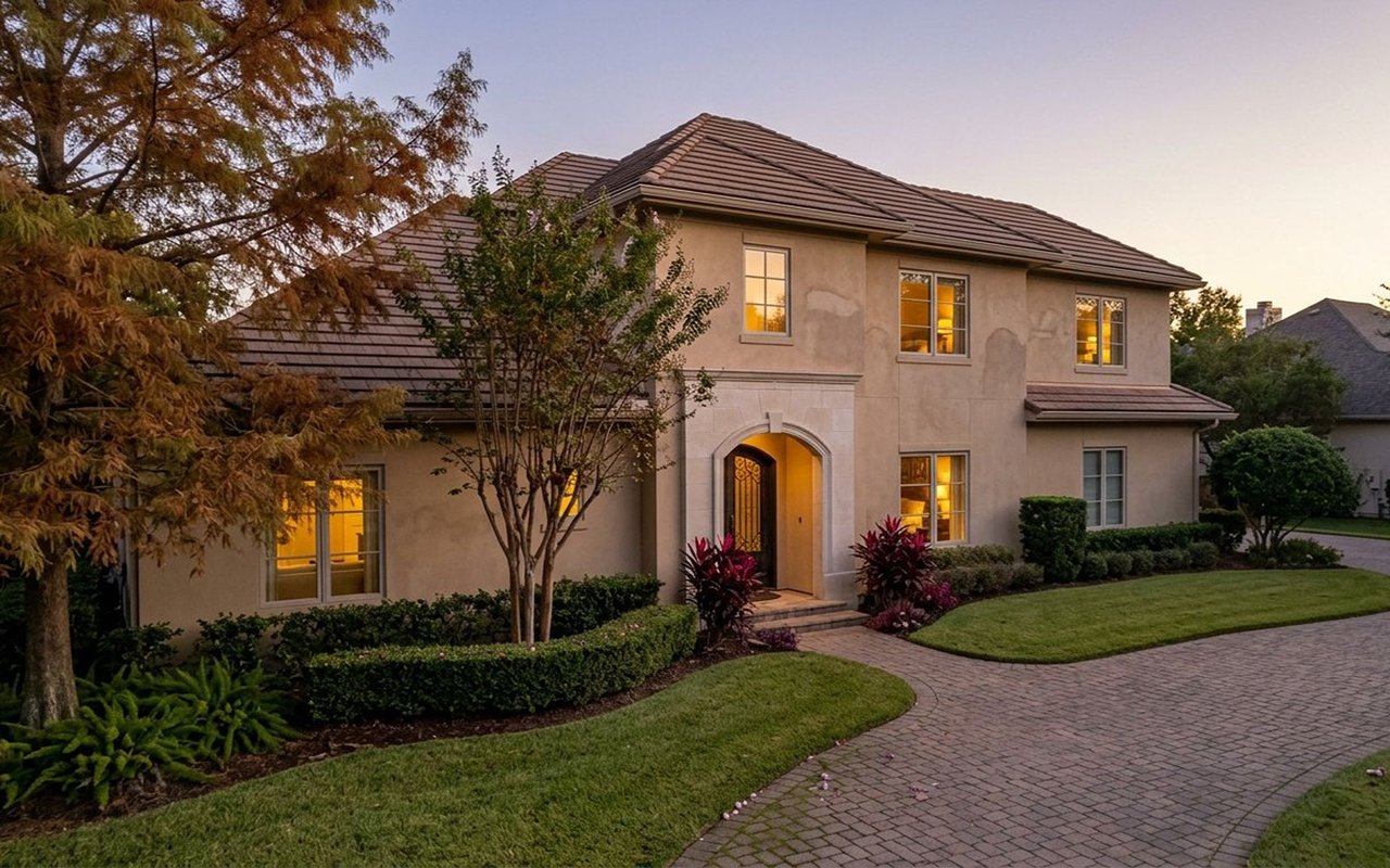 Two-story stucco home with an arched entry, lit windows, landscaped lawn, and curved brick driveway at dusk.