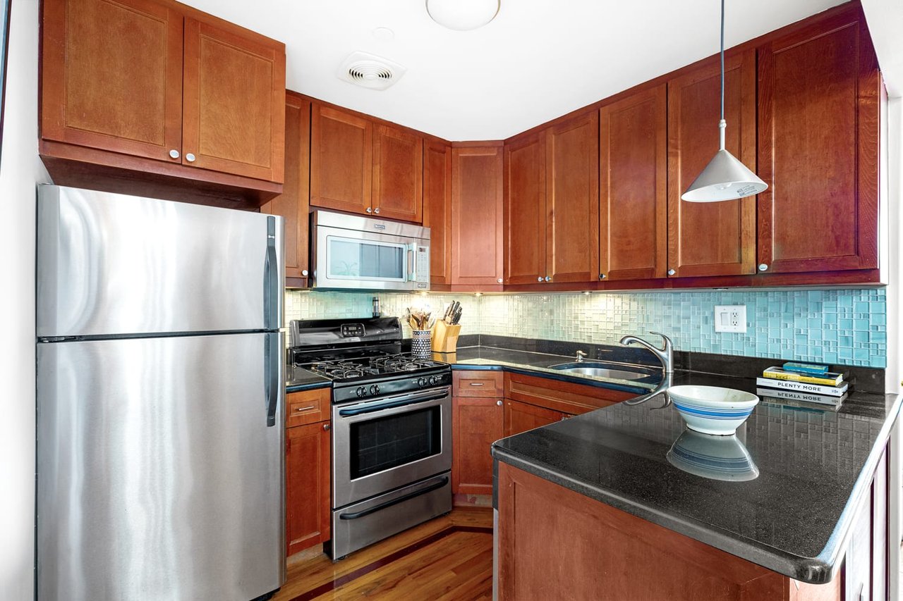 Apartment kitchen with reddish-brown cabinets, stainless steel appliances, turquoise tile backsplash, and a black countertop peninsula.