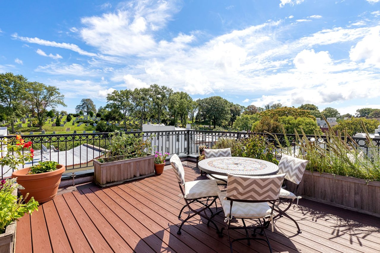 Rooftop terrace with outdoor dining set and planters overlooking a tree-filled cemetery.
