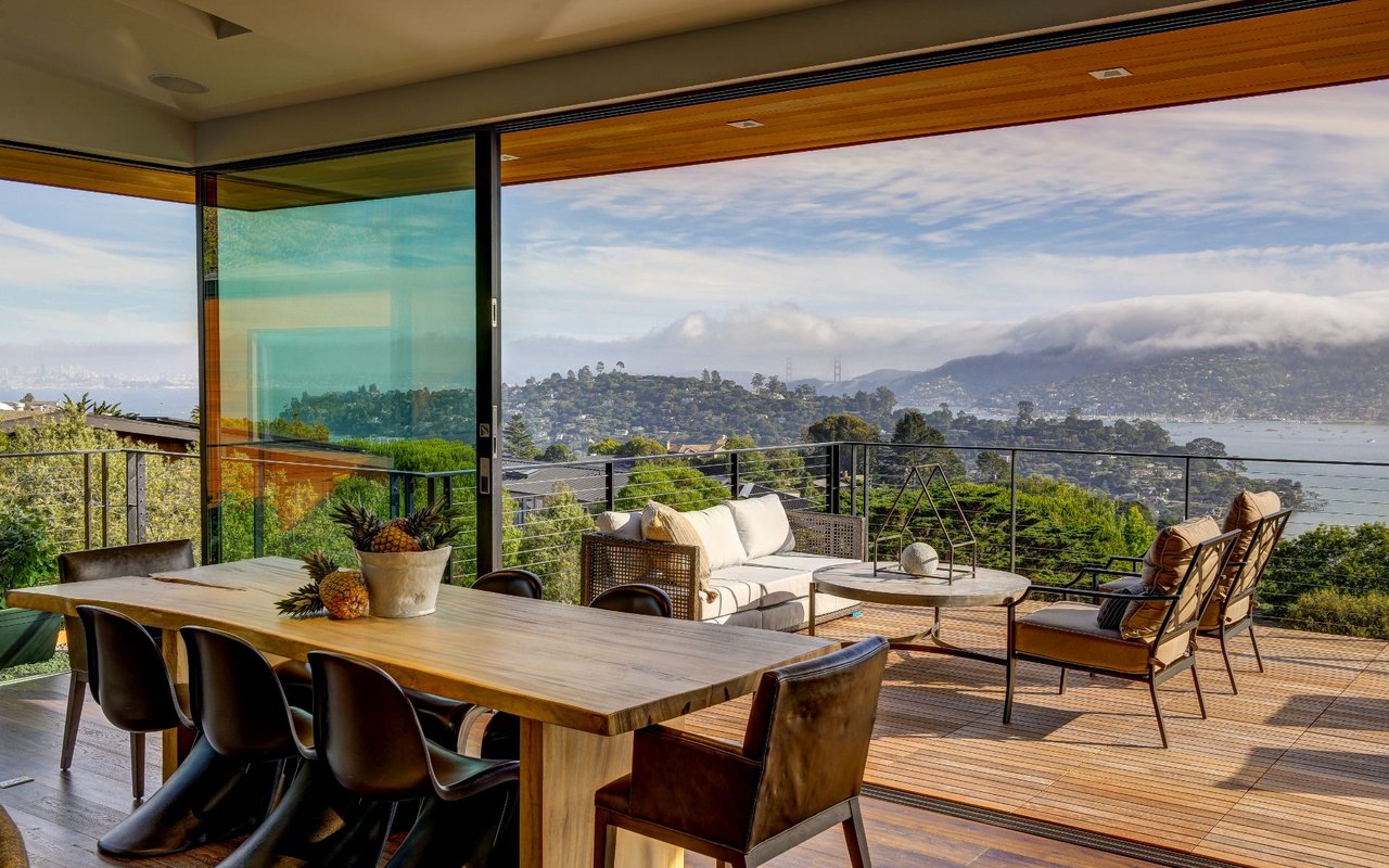 Furnished deck with a long dining table and lounge seating overlooking the bay and wooded hills.