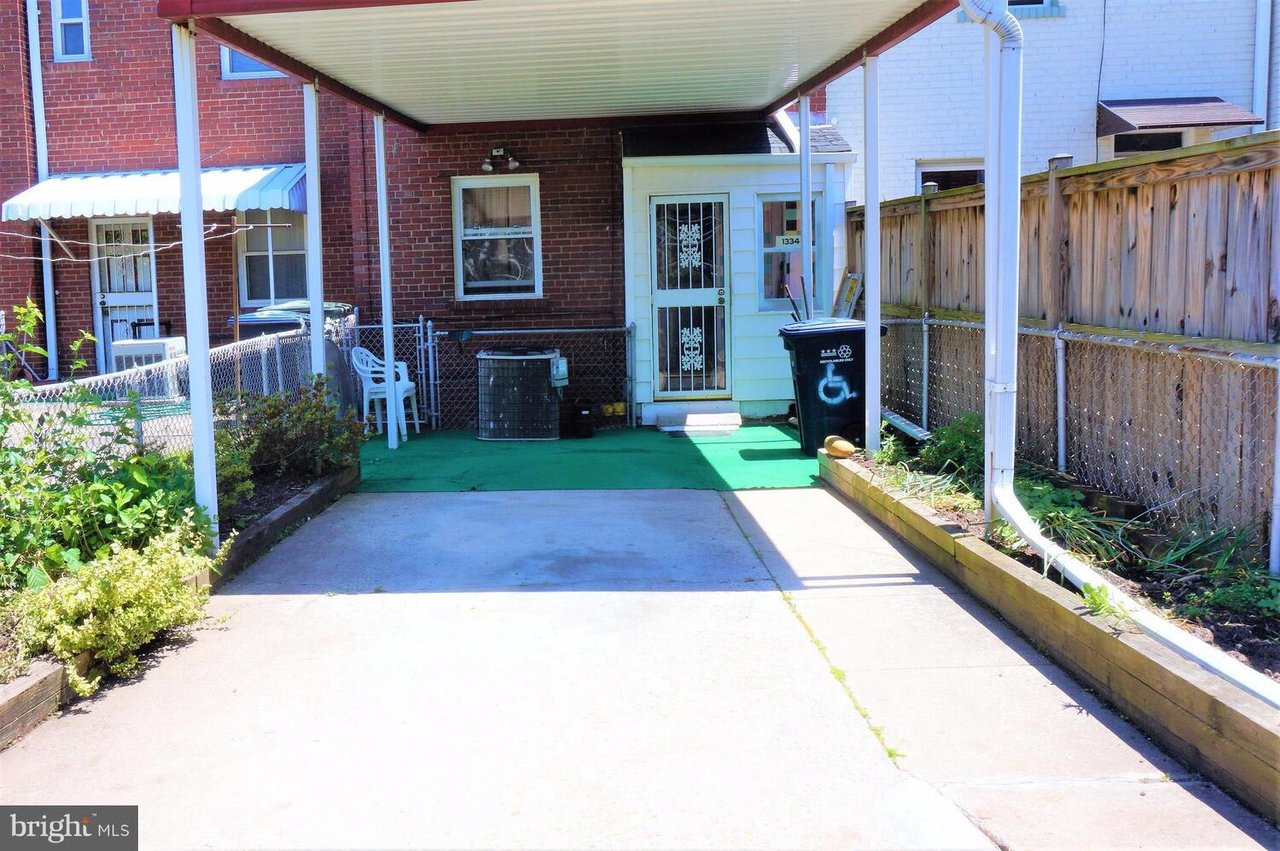 Covered driveway leading to a light-colored entry door, bordered by chain-link fencing and raised planting beds.