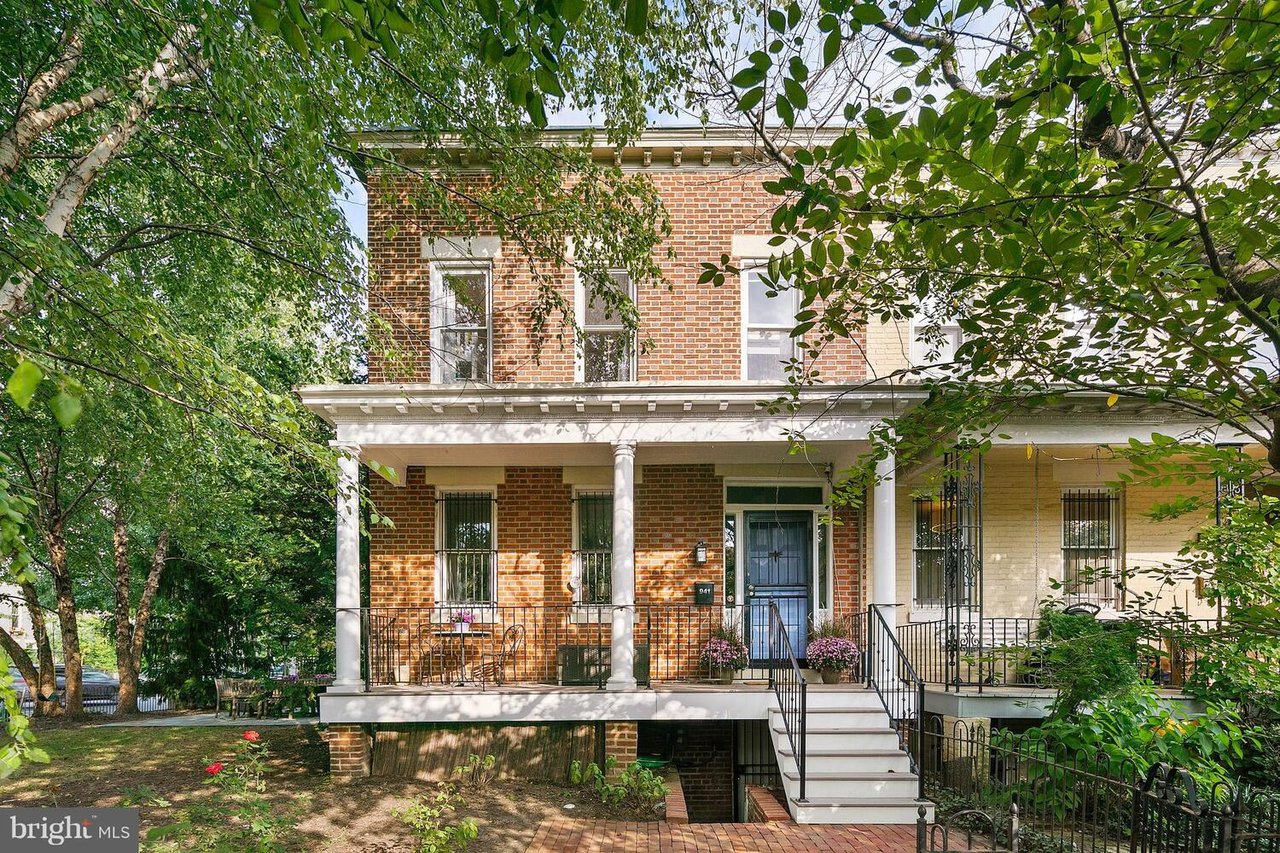 Red-brick rowhouse with a white-columned porch, blue door, and leafy trees framing the front yard.
