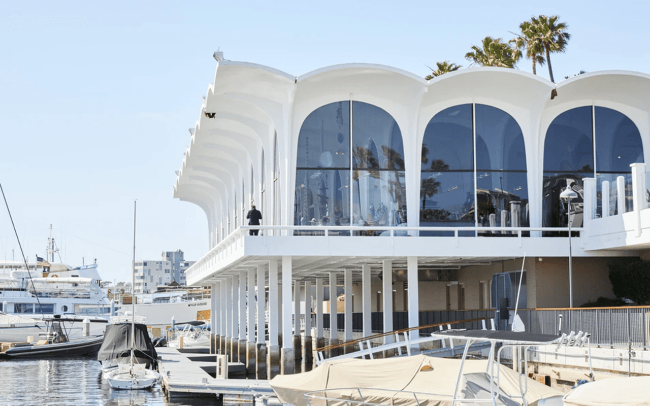 A futuristic white building with large arched windows overlooking a marina filled with boats and yachts on a sunny day.