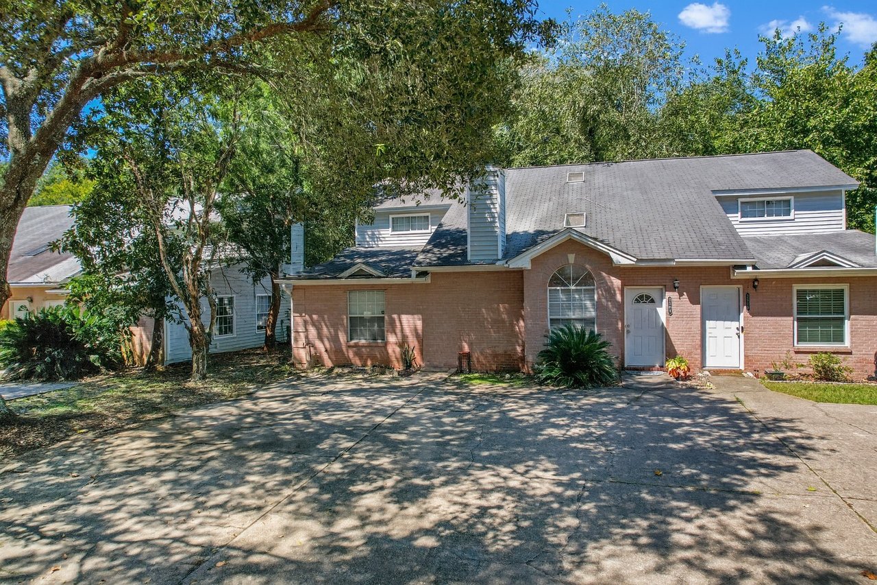 Front exterior view of a brick two-story townhome at 2506 Nugget Lane in Tallahassee, Florida. The home features a pitched roof with dormer windows, white trim, and two white front entry doors. A wide concrete driveway is shaded by mature trees, with some landscaping including small shrubs near the entrance.