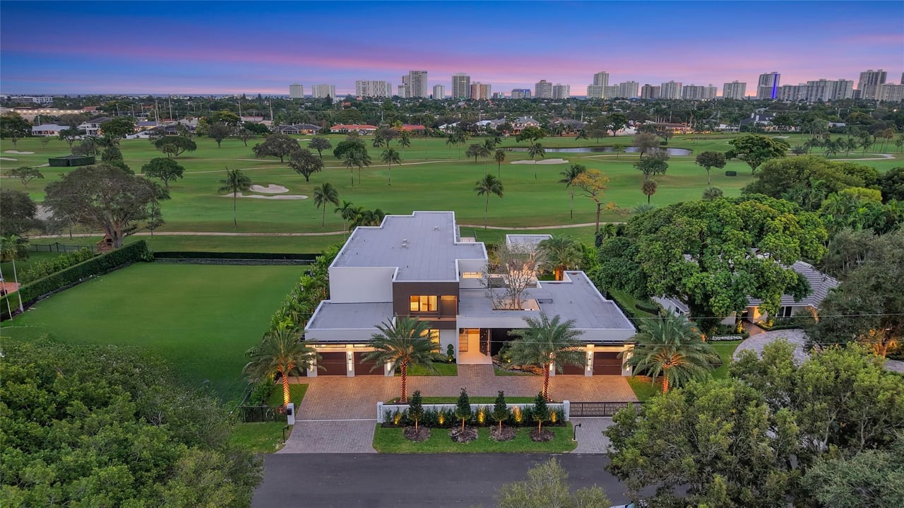 Modern home beside golf course fairways, with palms and a distant high-rise skyline at dusk.