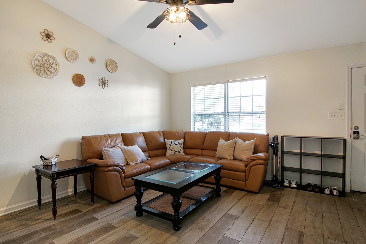 Cozy living room with a brown leather sectional, coffee table, and wall art. Warm lighting from a ceiling fan gives a welcoming, relaxed vibe.