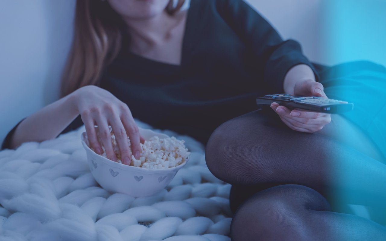 Person holding a remote and reaching for popcorn while seated on a sofa
