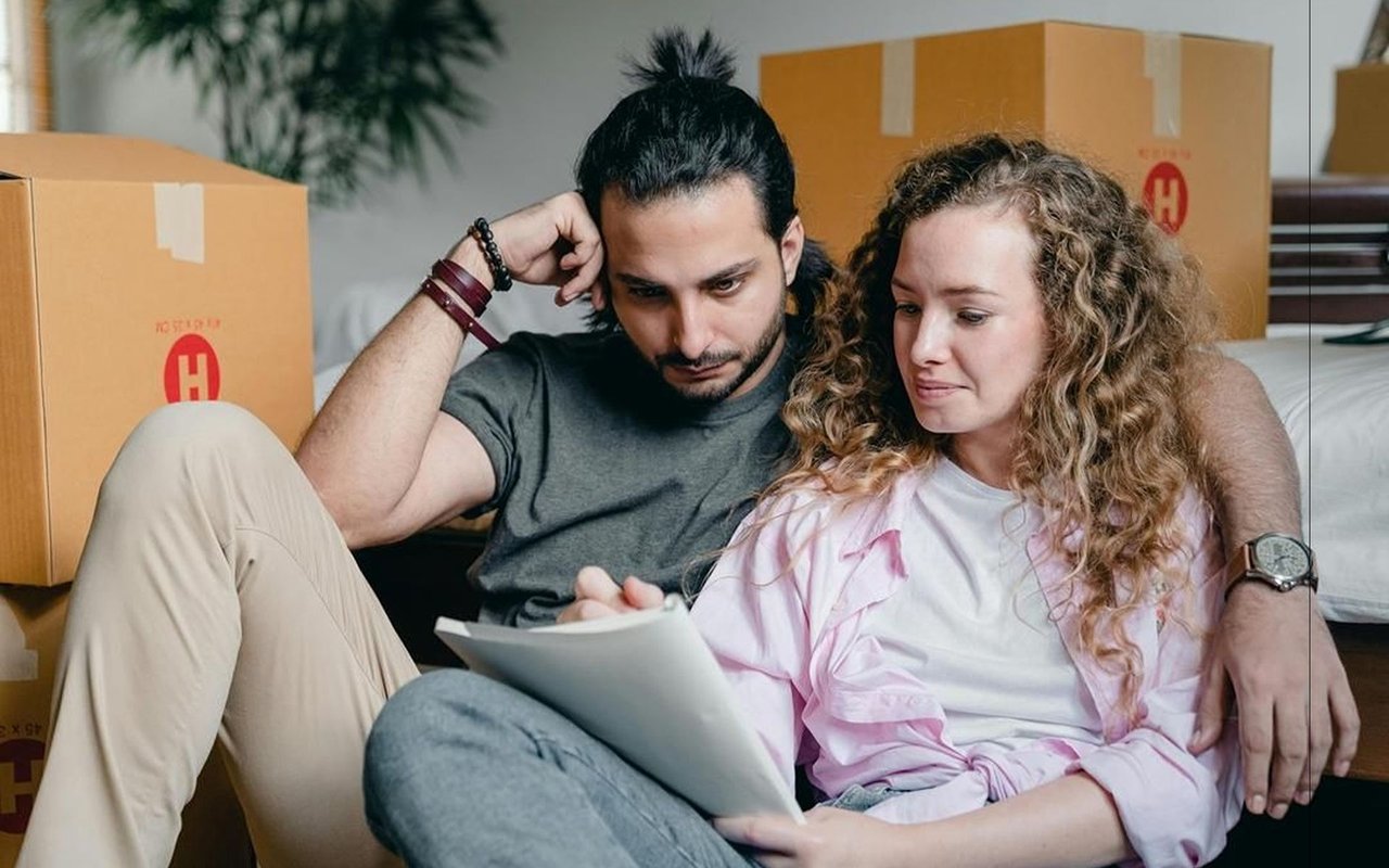Couple reviews a document together among moving boxes in a bedroom.