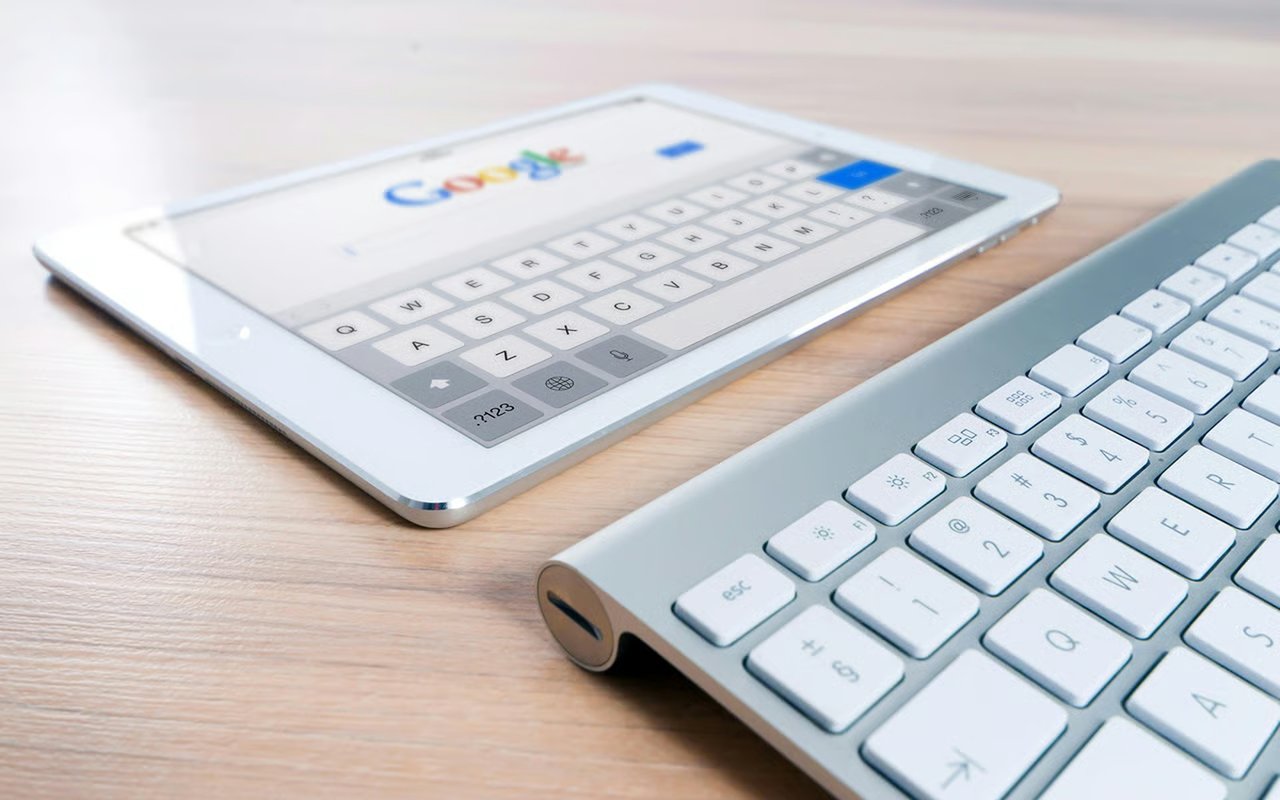 Tablet displaying Google beside a white computer keyboard on a wooden desk.