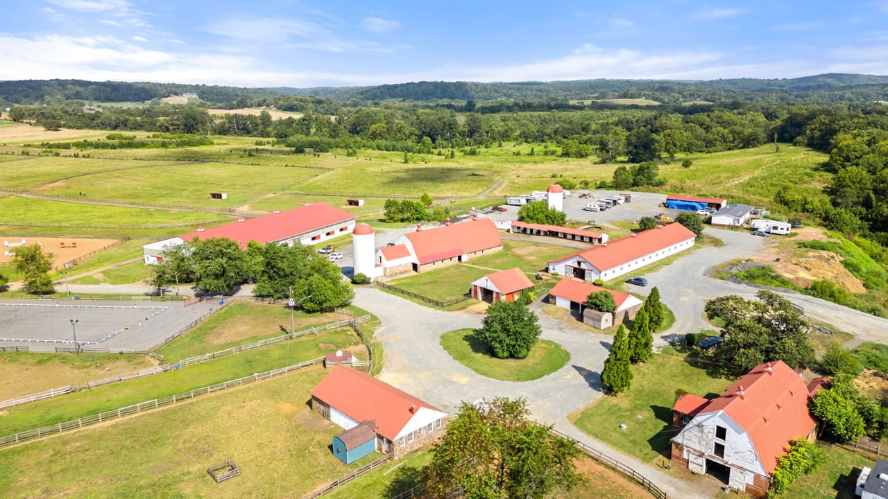 Aerial view of an equestrian property with red-roof barns, fenced paddocks, and rolling countryside.
