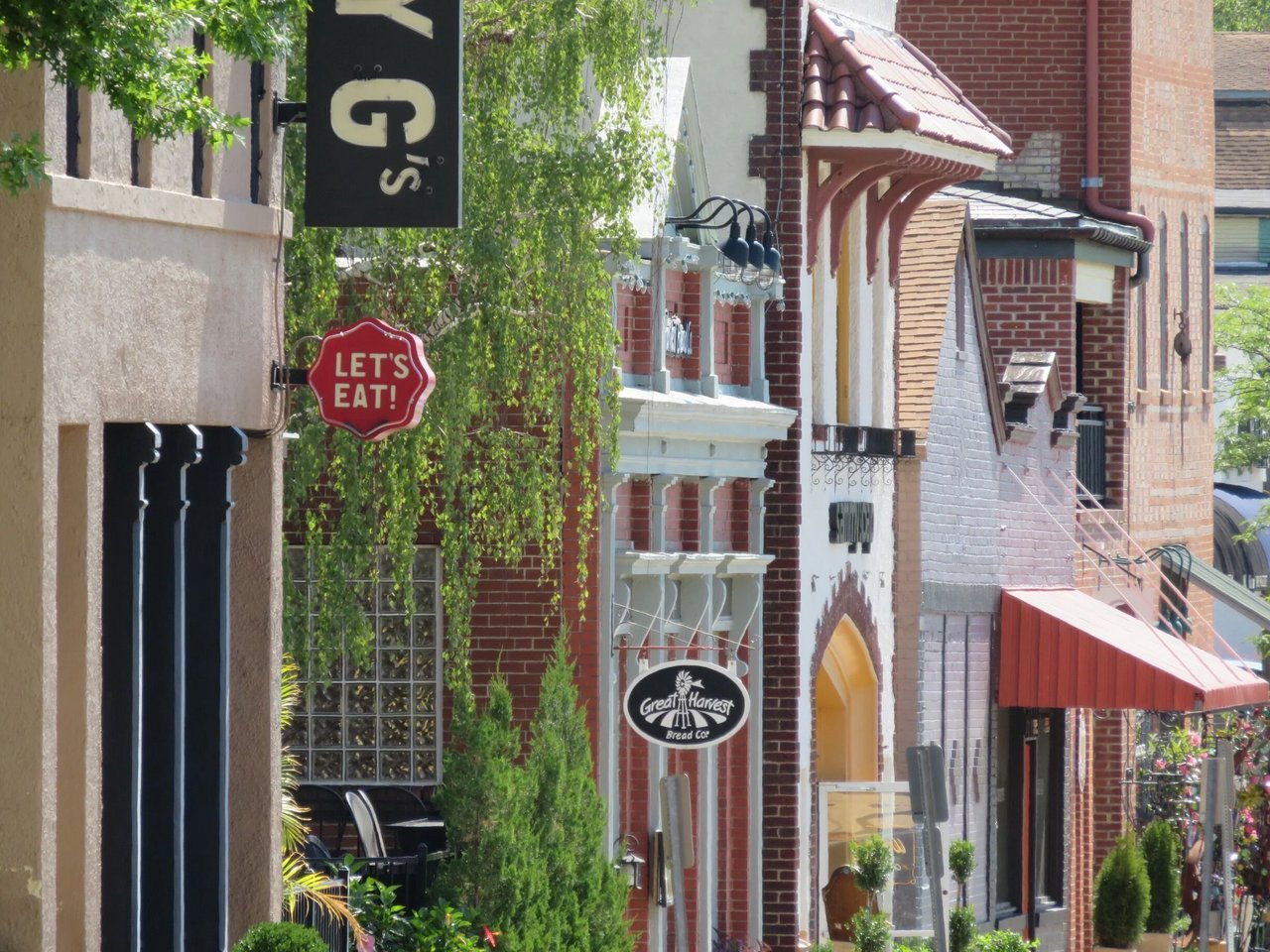 Commercial streetscape with brick storefronts, leafy trees, and a Great Harvest Bread Co. sign.
