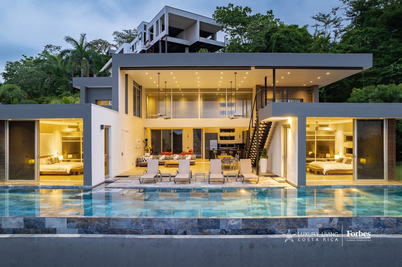 Modern tropical home with glass walls and a long pool, surrounded by dense greenery at dusk.
