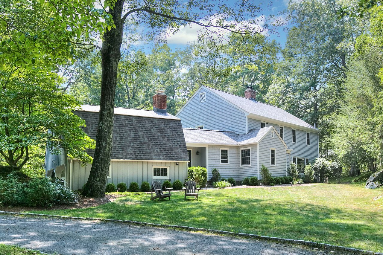 Light blue two-story house with gabled roofs and brick chimneys, framed by mature trees and a broad lawn.