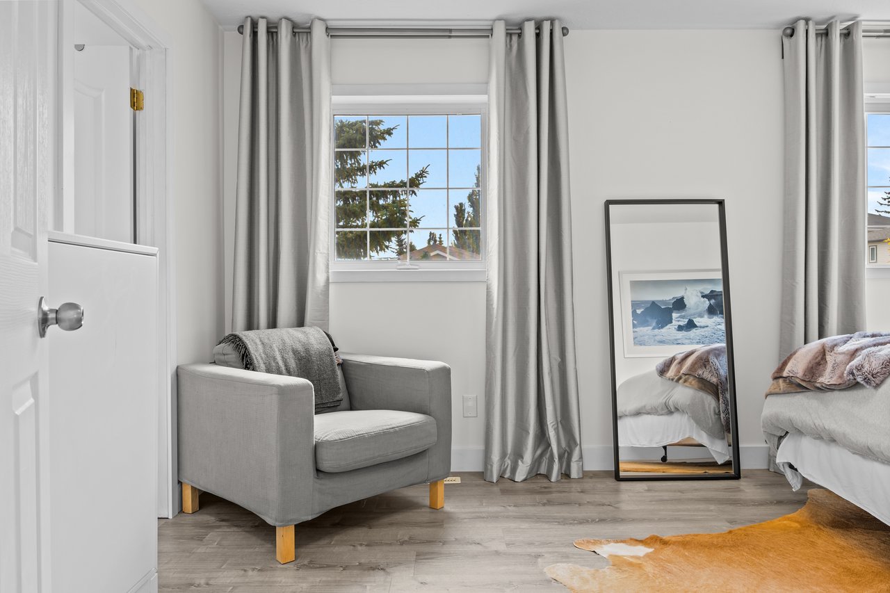 Bedroom with two beds, a gray armchair, and a full-length mirror beside curtained windows.