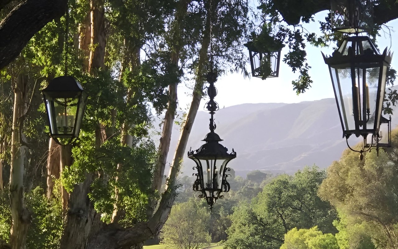 Ornate black lanterns hang among leafy trees with distant mountains behind them.