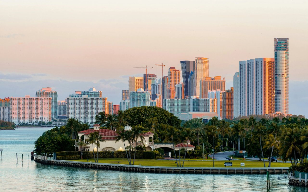 Waterfront estate and palms in front of a high-rise skyline across the water.
