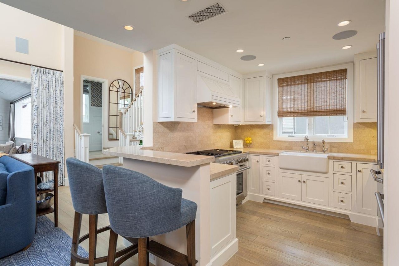  A modern kitchen with white cabinets, a farmhouse sink, and a counter with two blue bar stools separating it from the living room.