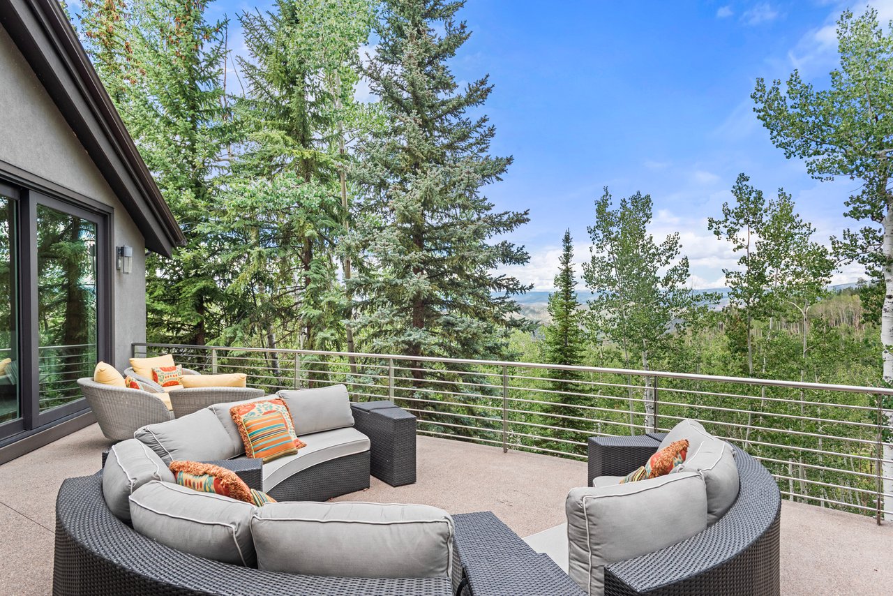 Mountain-home terrace with curved wicker seating overlooking a wooded hillside and distant mountains.