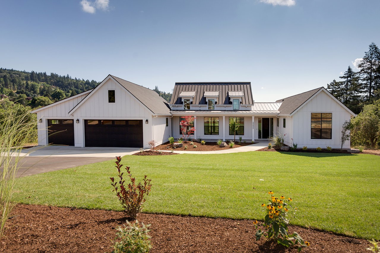 White modern farmhouse with a two-car garage, landscaped lawn, and wooded hills behind it.