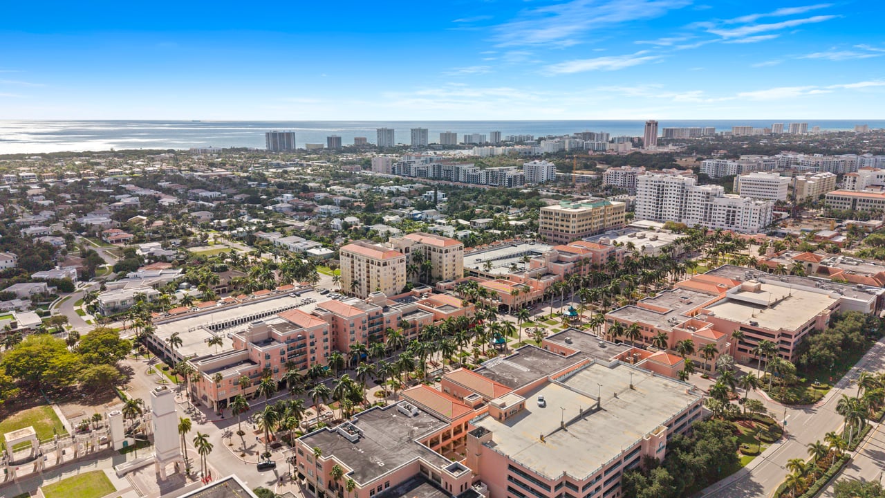 Aerial coastal cityscape with palm-lined streets, mixed-height buildings, and the ocean on the horizon.