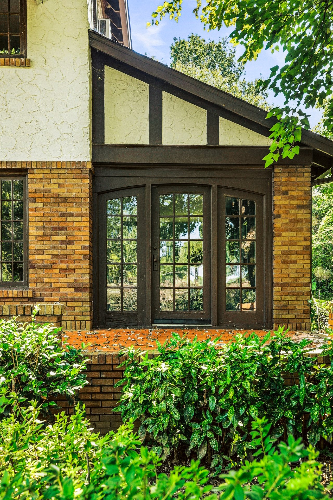 Brick house entrance with dark timber framing, glass double doors, and leafy plants.