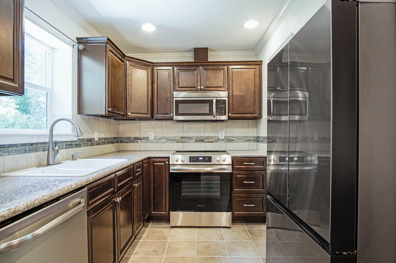 A modern kitchen with dark wood cabinets, granite countertops, stainless steel appliances, and a white sink. The room is well-lit with recessed lights.