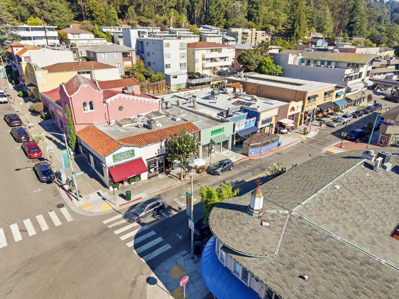 Aerial view of a commercial street with colorful storefronts and a wooded hillside beyond.
