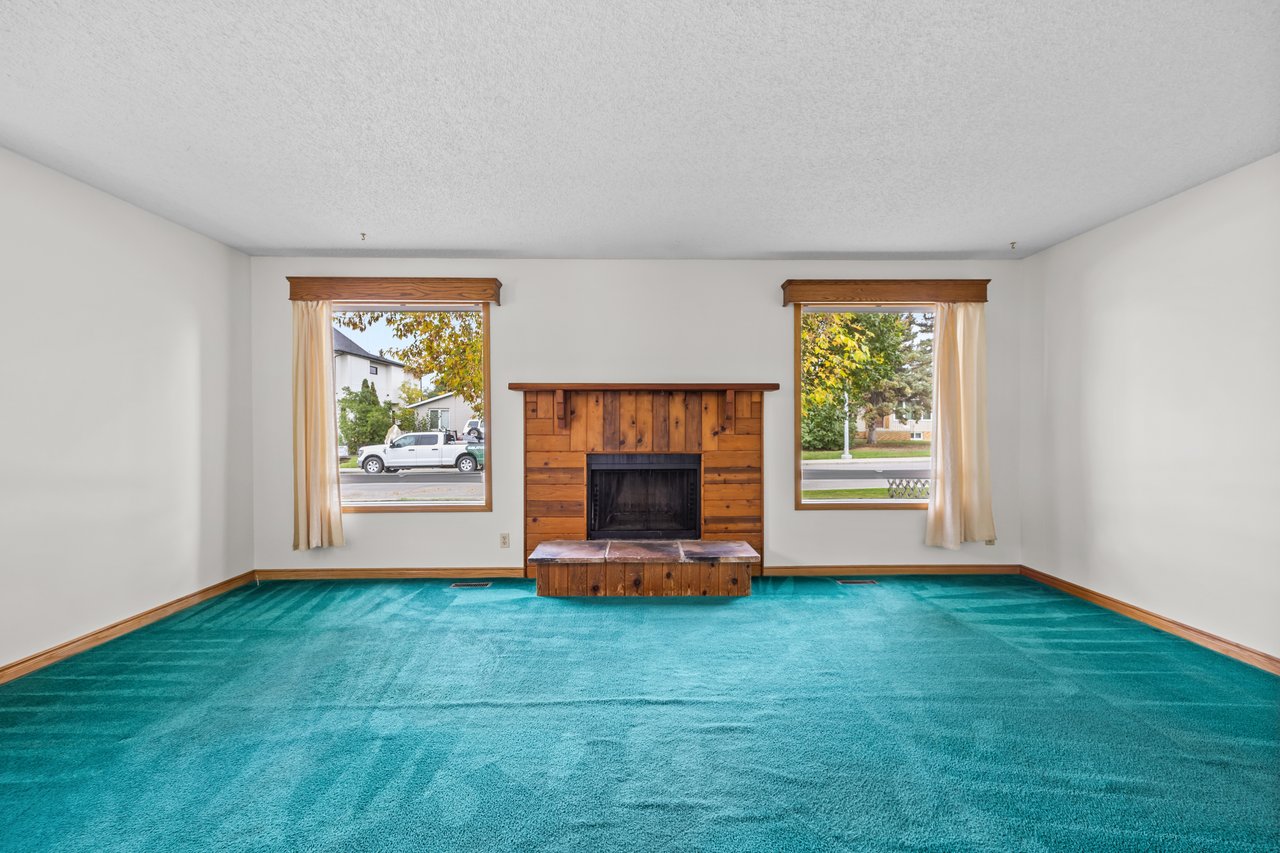 Empty living room with teal carpet and a wood-paneled fireplace between two windows.