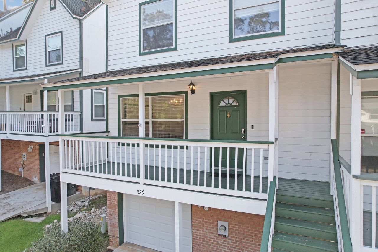 Front exterior of the townhome at 529 Collinsford Road, Tallahassee, Florida, featuring a covered porch with white railings, green trim, and a green front door. The home has white siding, a one-car garage with brick detailing, and a welcoming elevated entrance with steps leading to the porch.