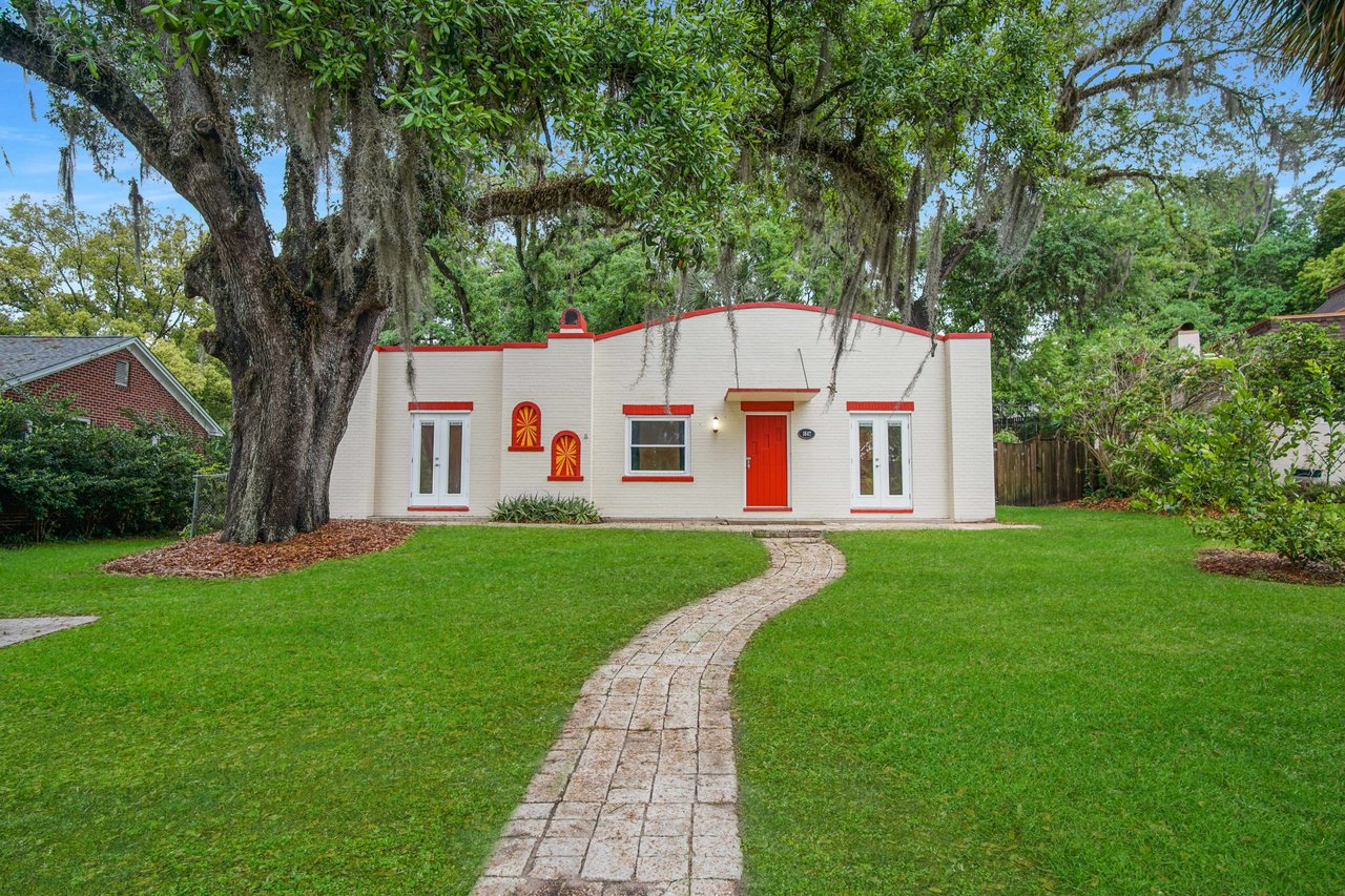 Solid brick and concrete light colored home with red trim, red front door, two glass French doors, pavered patio, and pavered walkway nestled under large oak trees.