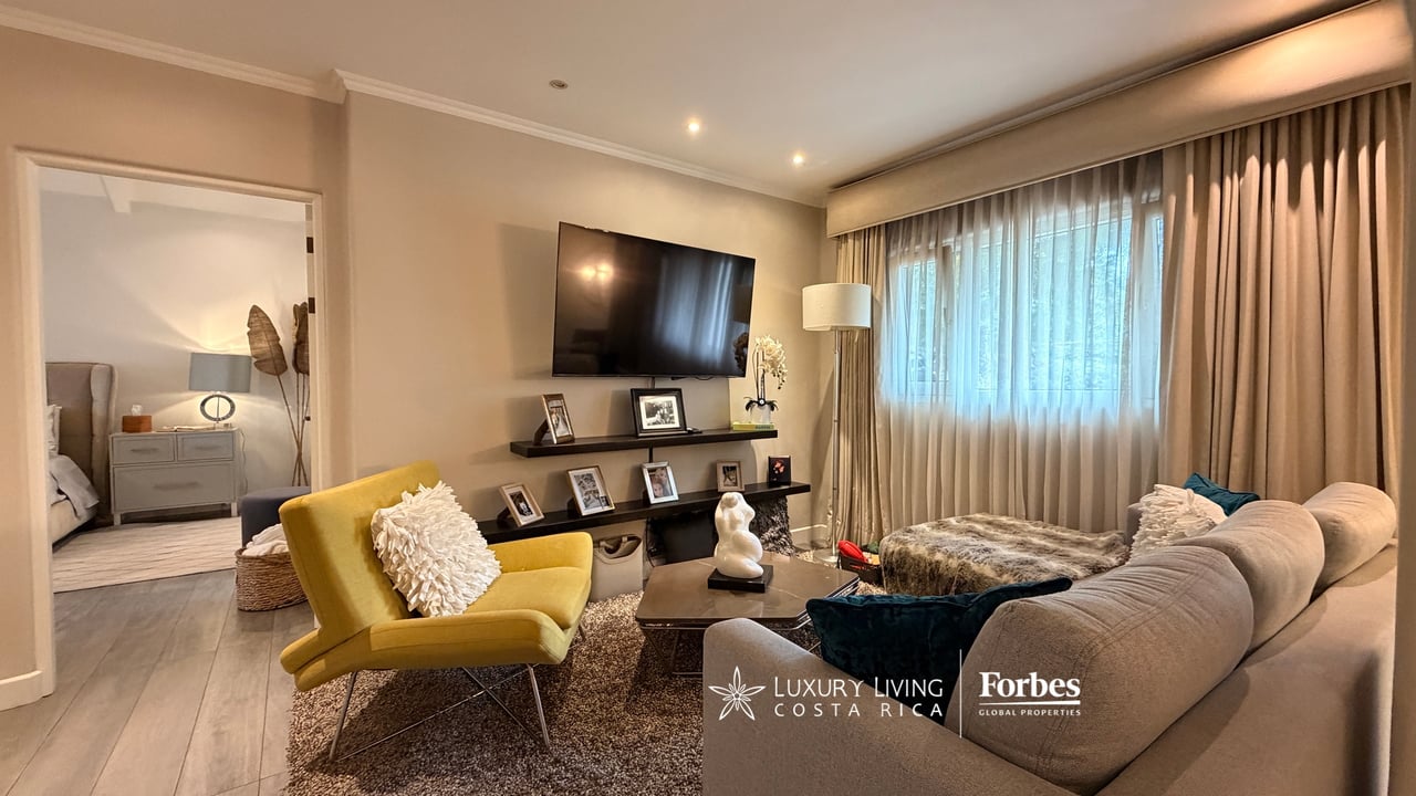 Furnished living room with a mustard-yellow chair, wall-mounted television, and adjoining bedroom doorway.