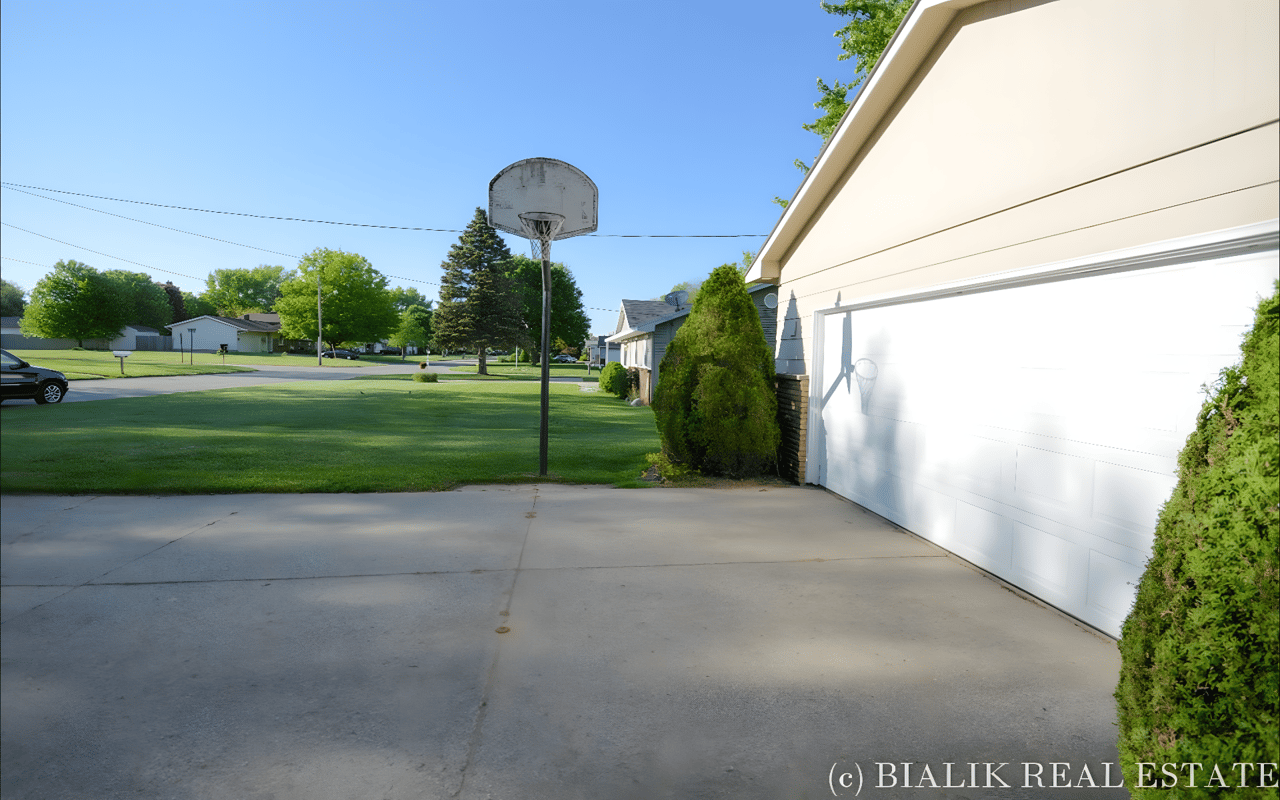 Residential driveway beside a white garage, with a basketball hoop and broad green lawn.